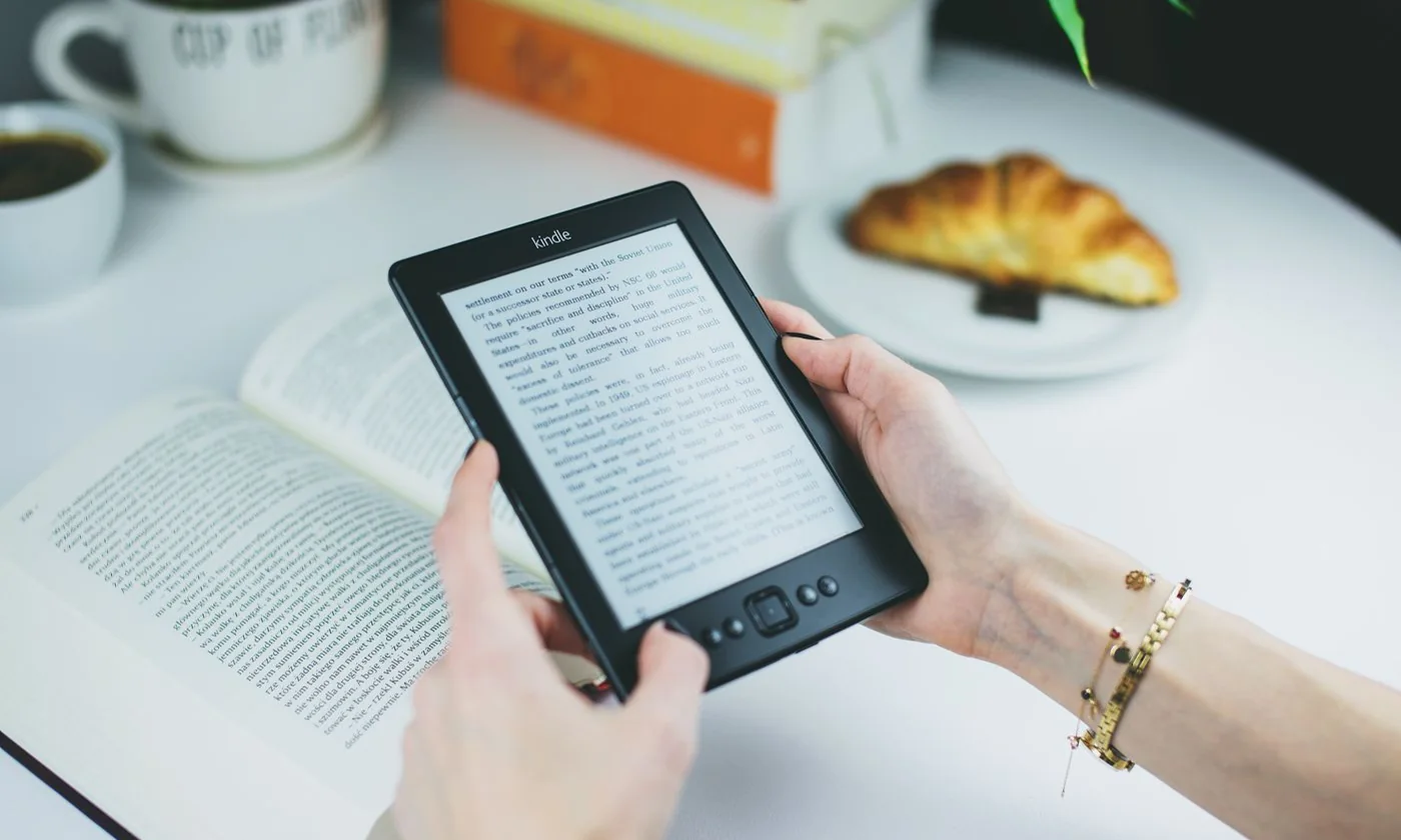 A person holding a Kindle e-reader at a table with a croissant and books
